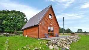 A wooden cabin with a sloped roof, a small porch with two chairs, and a stone wall in front. - Holiday Home in Swiecianowo near Lake Beach (Swiecianowo)