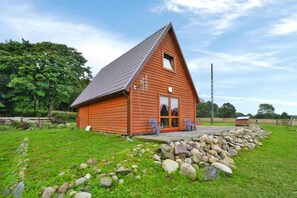 A wooden cabin with a sloped roof, a porch with two chairs, and a stone wall in front. - Holiday Home in Swiecianowo near Lake Beach (Swiecianowo)