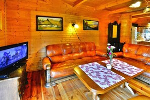 A cozy room with wooden walls, a leather sofa, a wooden coffee table with a floral table runner, and a television on a wooden cabinet. - Holiday Home in Swiecianowo near Lake Beach (Swiecianowo)