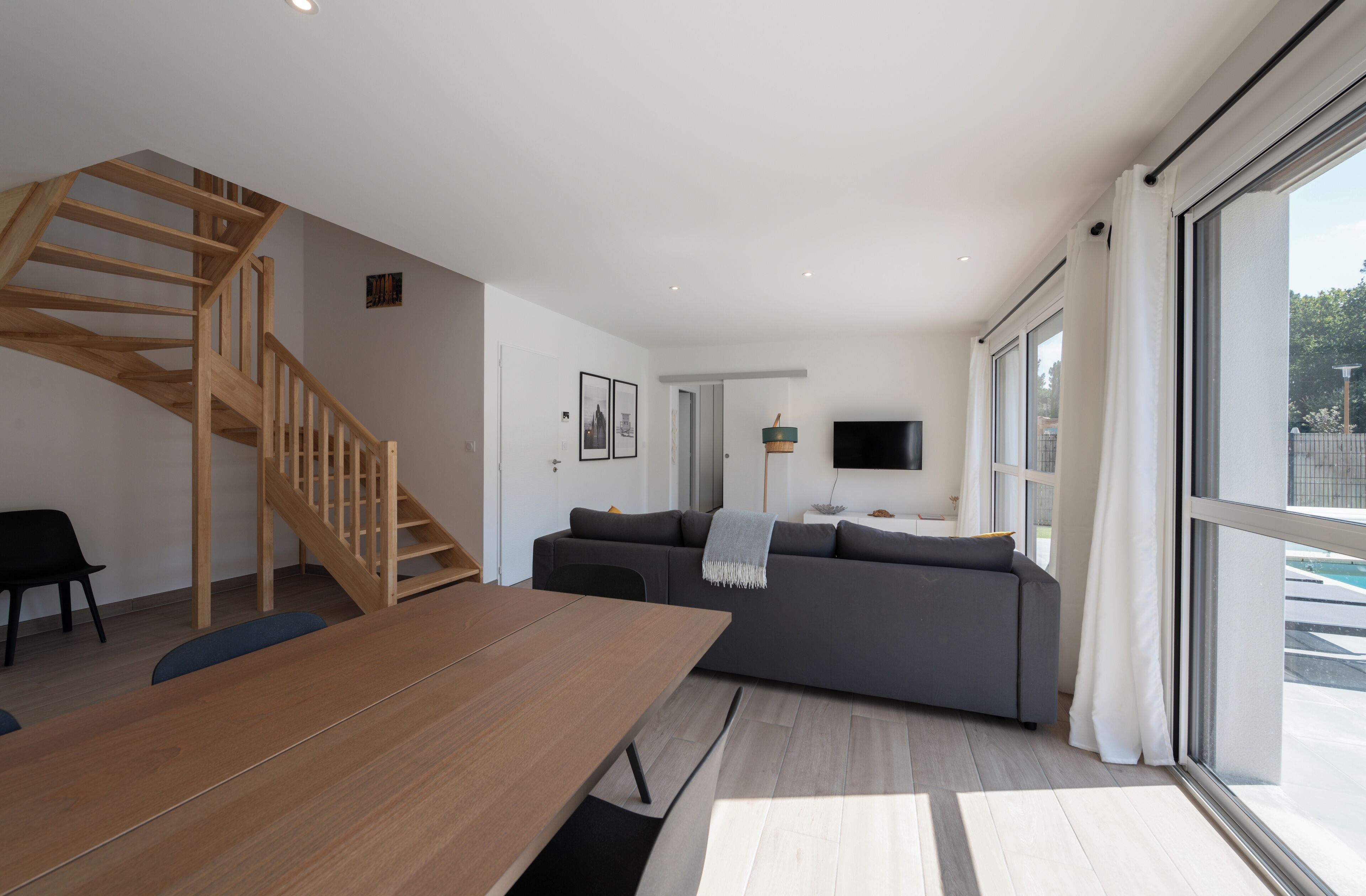 Un salon moderne avec un escalier en bois, un canapé gris, une table à manger en bois et de grandes fenêtres.
