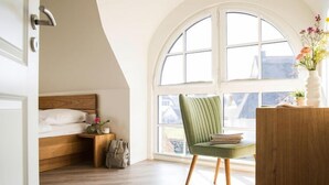 Une chambre avec un lit en bois, une chaise verte, un bureau en bois et une grande fenêtre en arc de cercle.