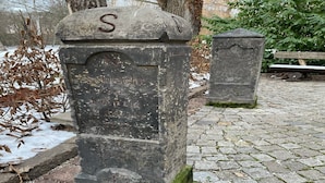 Una làpida de pedra desgastada amb inscripcions, situada sobre un camí empedrat i amb una banca al fons.