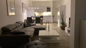 Un salon moderne avec un canapé d’angle, une table basse et un coin repas comprenant une table et des chaises. On aperçoit, au fond, une cuisine avec des meubles blancs et des appareils électroménagers.