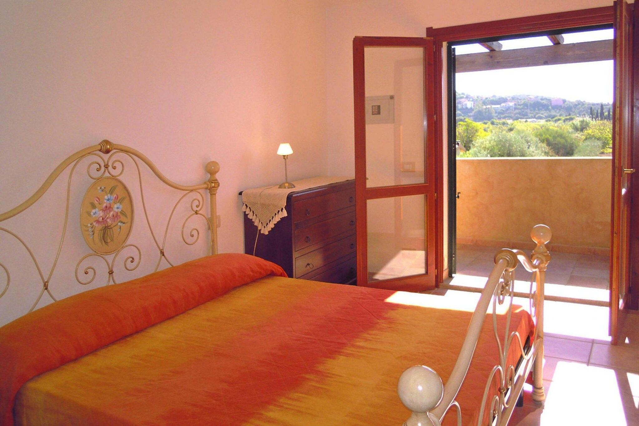 A bedroom with a bed, a dresser, a lamp, and a door leading to a balcony with a view.