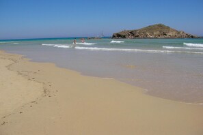 A sandy beach with gentle waves, a rocky outcrop in the distance, and a clear blue sky. - Apartment in a seaside resort (Domus De Maria / Chia)