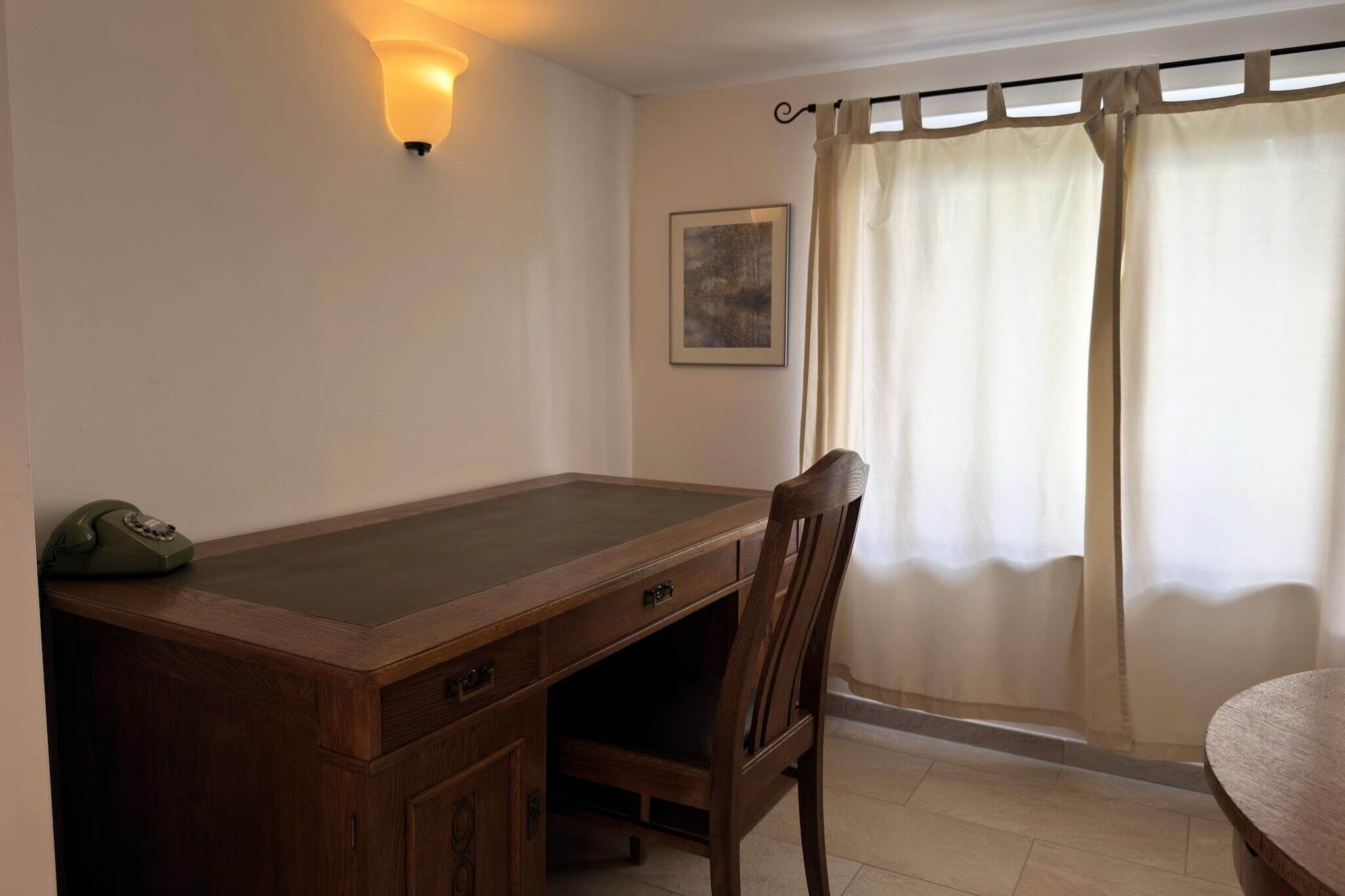 A room with a wooden desk, a chair, a telephone, a framed picture on the wall, and a window with curtains.