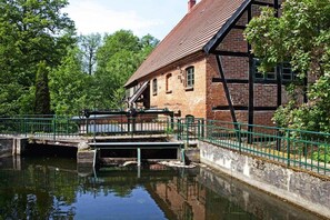 Un bâtiment traditionnel en briques, doté d’un toit à forte pente, d’un petit pont enjambant un canal et d’une rambarde métallique verte le long des berges du canal.