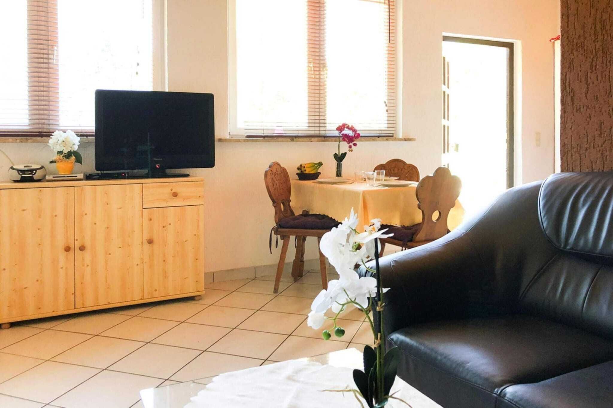 Una sala de estar con un sofá de cuero negro, un mueble de madera y un televisor. Hay una mesa de comedor con sillas y un jarrón de flores.