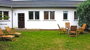 Une maison à l’extérieur blanc, avec un toit en tuiles et un petit jardin devant, composé d’une pelouse et de chaises en bois.
