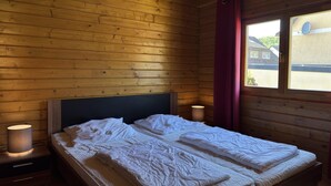 Une chambre dans un chalet en bois, avec un lit, deux lampes de chevet et une fenêtre donnant sur des maisons.