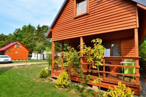 A wooden cabin with a porch, surrounded by greenery and a car in the background.
