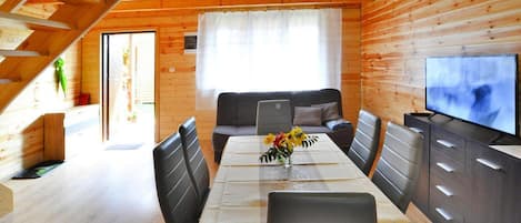 A wooden cabin interior with a dining table, chairs, a sofa, a television, and a window with curtains.