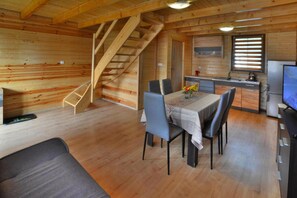 A wooden cabin interior with a dining table, chairs, a sofa, a kitchen area, and a staircase.