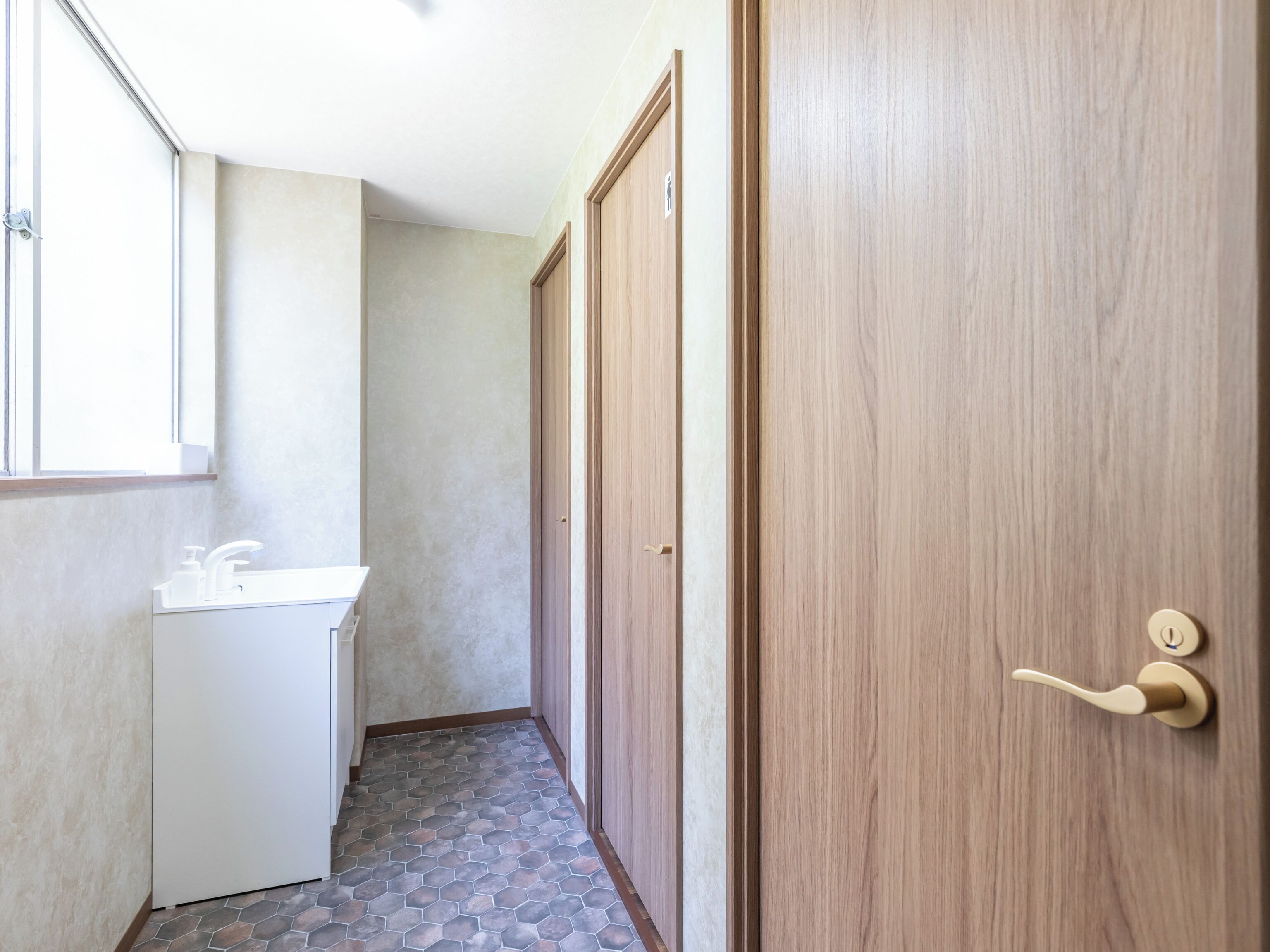 Ein schmaler Flur mit einem Badezimmer links, das ein Waschbecken und eine Toilette zeigt. Der Boden ist mit sechseckigen Fliesen belegt, und rechts befinden sich zwei Holztüren.