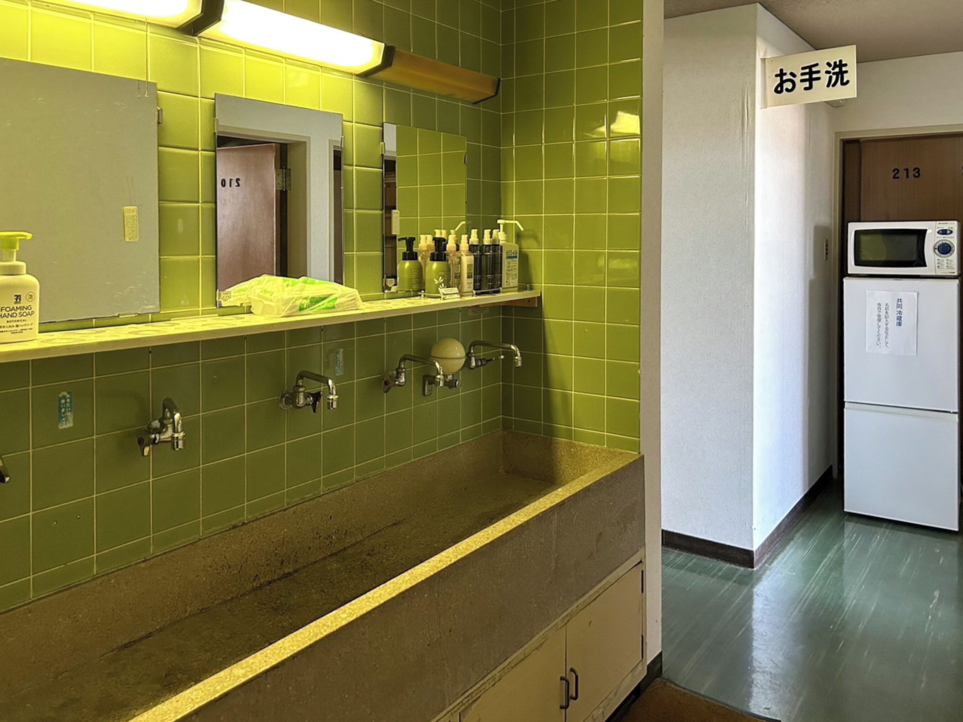 A bathroom with green tiles, a sink, and a mirror. There is a shelf with toiletries and a sign above the sink that says "hand wash".