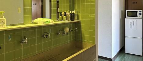 A bathroom with green tiles, a sink, and a mirror. There is a shelf with toiletries and a sign above the sink that says "hand wash".