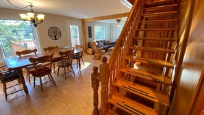 A spacious living area with a wooden staircase, a dining table with chairs, and a large window providing natural light. - Country Chalet with Lakefront (Chertsey)