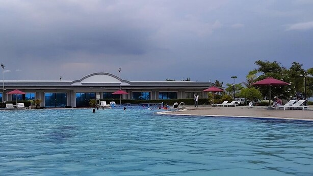 En swimmingpool med liggestole og parasoller, en bygning med store vinduer, og en overskyet himmel.