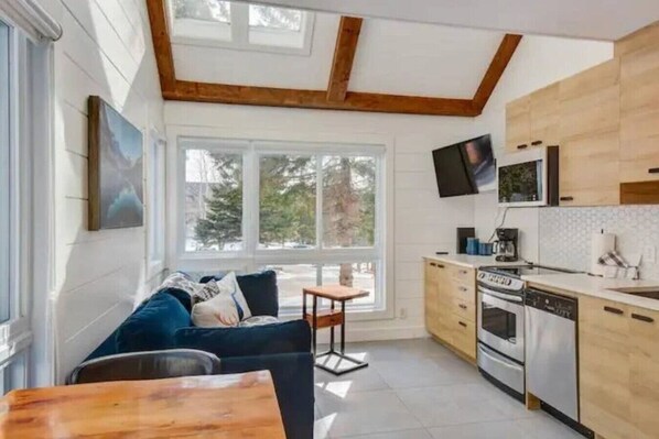 Eine moderne Küche mit Holzschränken, einem eingebauten Ofen und einem Geschirrspüler. Ein Esstisch mit Stühlen, eine Couch und ein an der Wand befestigter Fernseher sind ebenfalls vorhanden. Große Fenster sorgen für natürliches Licht und bieten Blick auf Bäume.