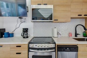 A kitchen with a microwave, stove, and dishwasher. There are wooden cabinets, a tiled backsplash, and a countertop with a coffee maker and a roll of paper towels. - Modern Cottage in the Tremblant Ski Village (Mont-Tremblant)