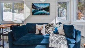 Una habitación acogedora con un sofá azul, un cuadro de un lago de montaña y vistas a un paisaje nevado a través de las ventanas.
