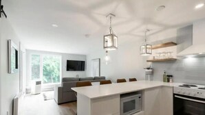 Une cuisine moderne dotée d’un îlot central, d’un four encastré et d’appareils électroménagers en acier inoxydable. On y trouve un coin repas avec un canapé et des chaises, ainsi qu’une grande fenêtre qui laisse entrer la lumière naturelle.