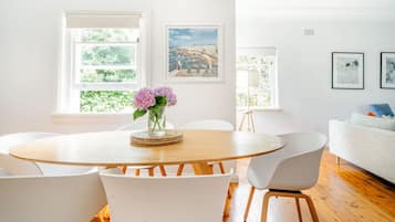 A modern living room with a round wooden table, white chairs, and a white sofa. There are two windows with views of greenery and a cityscape.