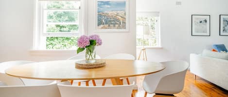 A modern living room with a round wooden table, white chairs, and a white sofa. There are two windows with views of greenery and a cityscape.