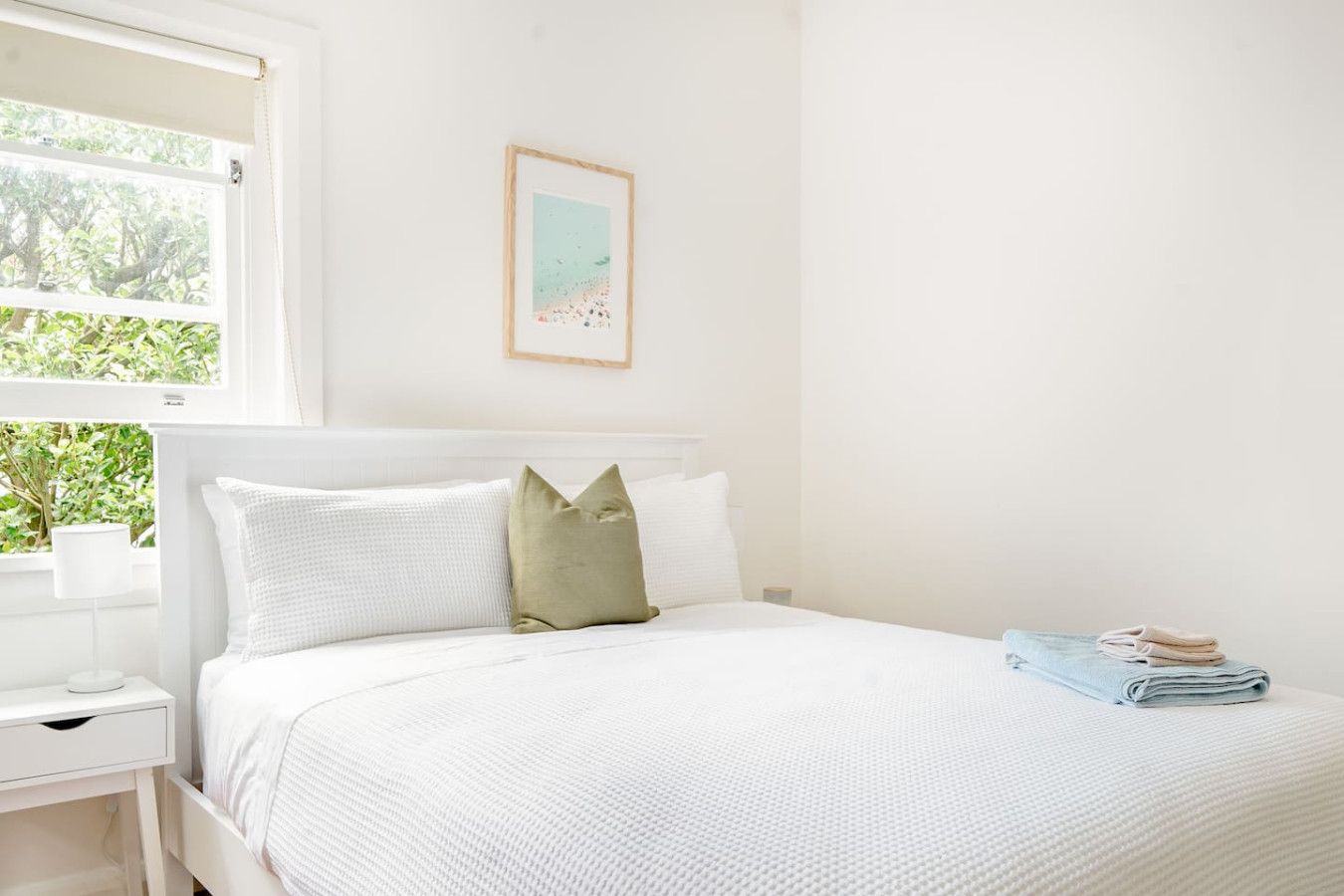 A white bedroom with a bed, a nightstand, a window with a view of greenery, and a framed picture on the wall.
