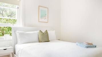 A white bedroom with a bed, a nightstand, a window with a view of greenery, and a framed picture on the wall.