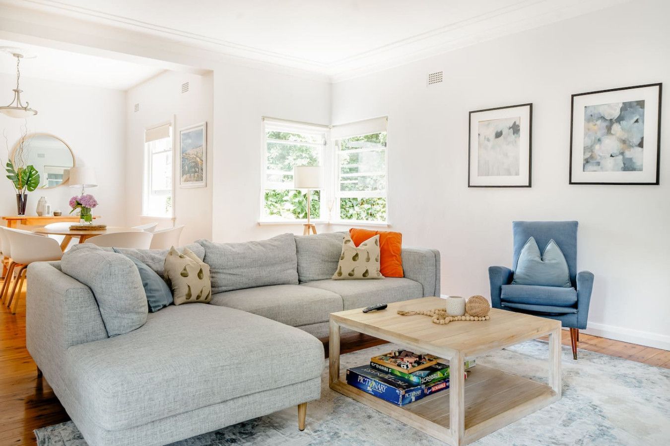 A modern living room with a grey sectional sofa, a coffee table, and a blue armchair. There are two windows with views of greenery, a dining area with a white table and chairs, and a wall with framed artwork.