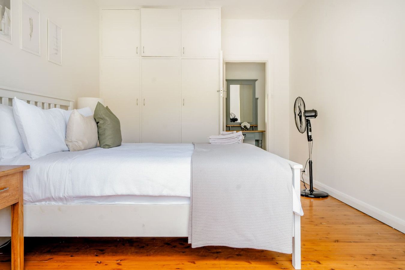 A bedroom with a bed, a bedside table, a mirror, and a standing fan.
