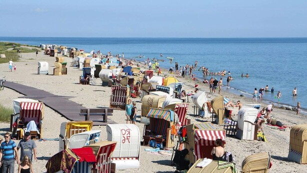 Una playa con numerosas sillas y sombrillas, un paseo marítimo de madera, y gente disfrutando del mar.