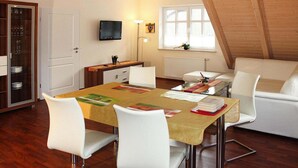 Un comedor con una mesa dispuesta para cuatro personas, sillas blancas, un televisor montado en la pared, un ventanal con vistas al exterior y un techo de madera.