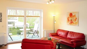 Una sala de estar con un sofá rojo, una mesa con un florero, una lámpara de pie y un ventanal corredizo que da a un área exterior con una mesa y sillas.