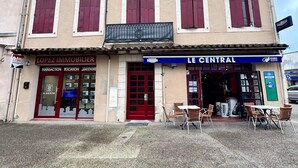 Un bâtiment avec une porte rouge et de nombreuses fenêtres dotées de volets. On y voit une enseigne indiquant « LOPEZ IMMOBILIER » et une autre indiquant « LE CENTRAL ».