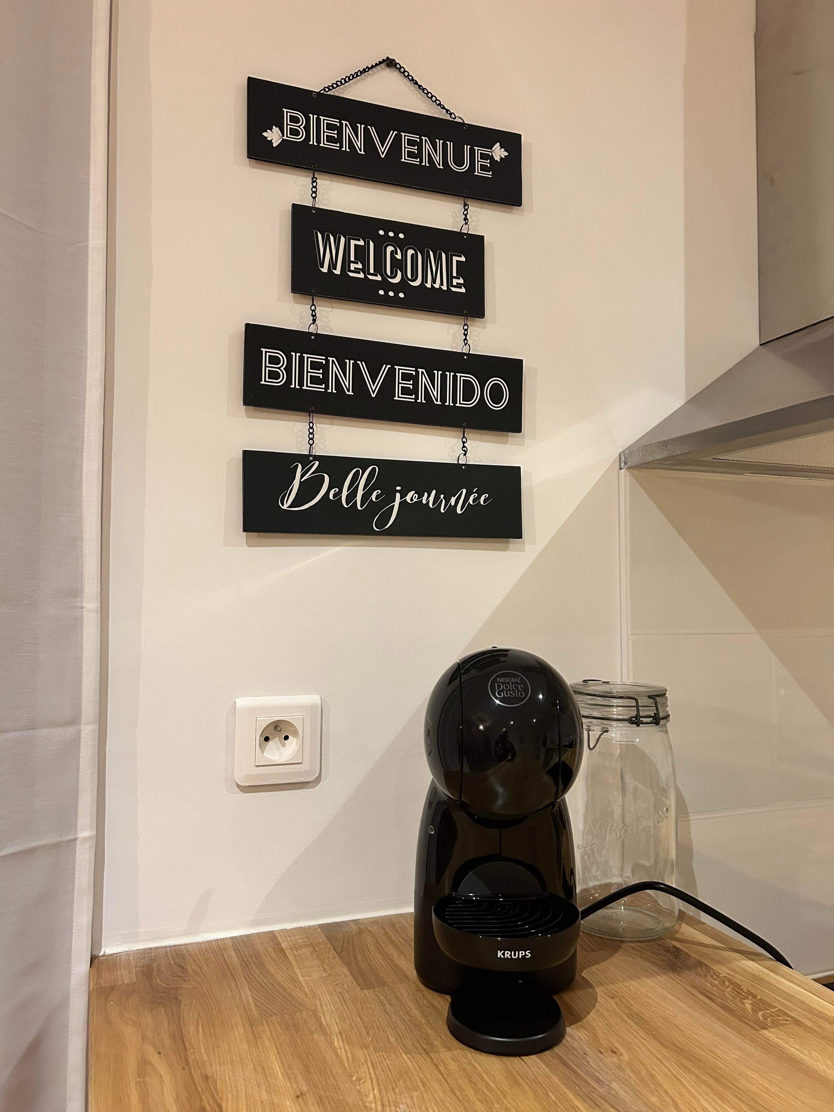 Eine Kaffeemaschine auf einer Holzauskładung, über ihr eine Wandtafel mit den Aufschriften „BIENVENUE“, „WELCOME“, „BIENVENIDO“ und „Belle journée“.