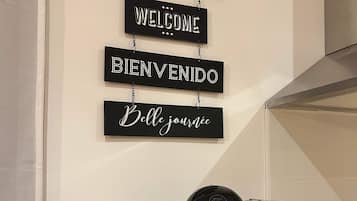 Eine Kaffeemaschine auf einer Holzauskładung, über ihr eine Wandtafel mit den Aufschriften „BIENVENUE“, „WELCOME“, „BIENVENIDO“ und „Belle journée“.