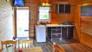 A compact kitchen with a refrigerator, stove, and cabinets. There is a window with curtains and a television mounted on the wall.
