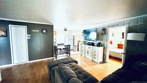 Ein Wohnzimmer mit grauem Sofa, einem Fernseher und einer Küche im Hintergrund.