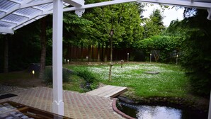 Een overdekt terras met een pad van bakstenen dat naar een vijver leidt, omringd door weelderige begroeiing en een houten terras.