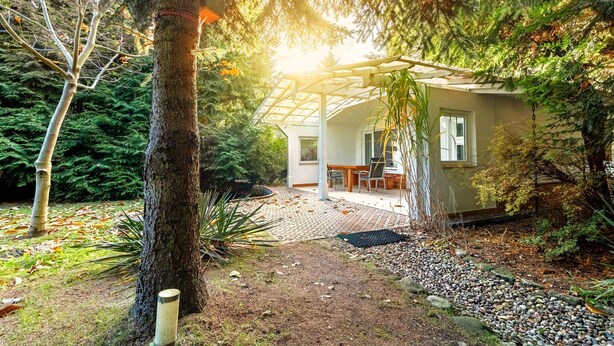 Een huis met een overdekte patio, omringd door weelderige begroeiing en een grindpad dat naar de ingang leidt.