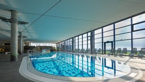 An indoor swimming pool with clear blue water, surrounded by large windows offering a view of the city skyline. - Ferienhaus in Damp mit Meeresstrand (Damp)
