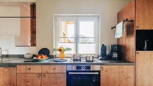 A modern kitchen with wooden cabinets, a built-in oven, a sink, and a window with a view of the outside. - Doppelhaushälften, Torfhaus (Torfhaus)