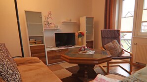 A living room with a sofa, a coffee table, a television, and a chair. There are bookshelves and a window with curtains. - Ferienhäuser am Teich, Zinnowitz (Zinnowitz)