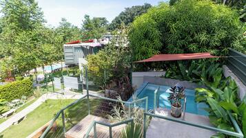 A rooftop pool area with a wooden deck, surrounded by lush greenery and a view of neighboring buildings.