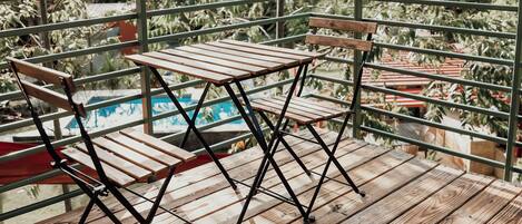 A wooden deck with a table and chairs, overlooking a pool and greenery.