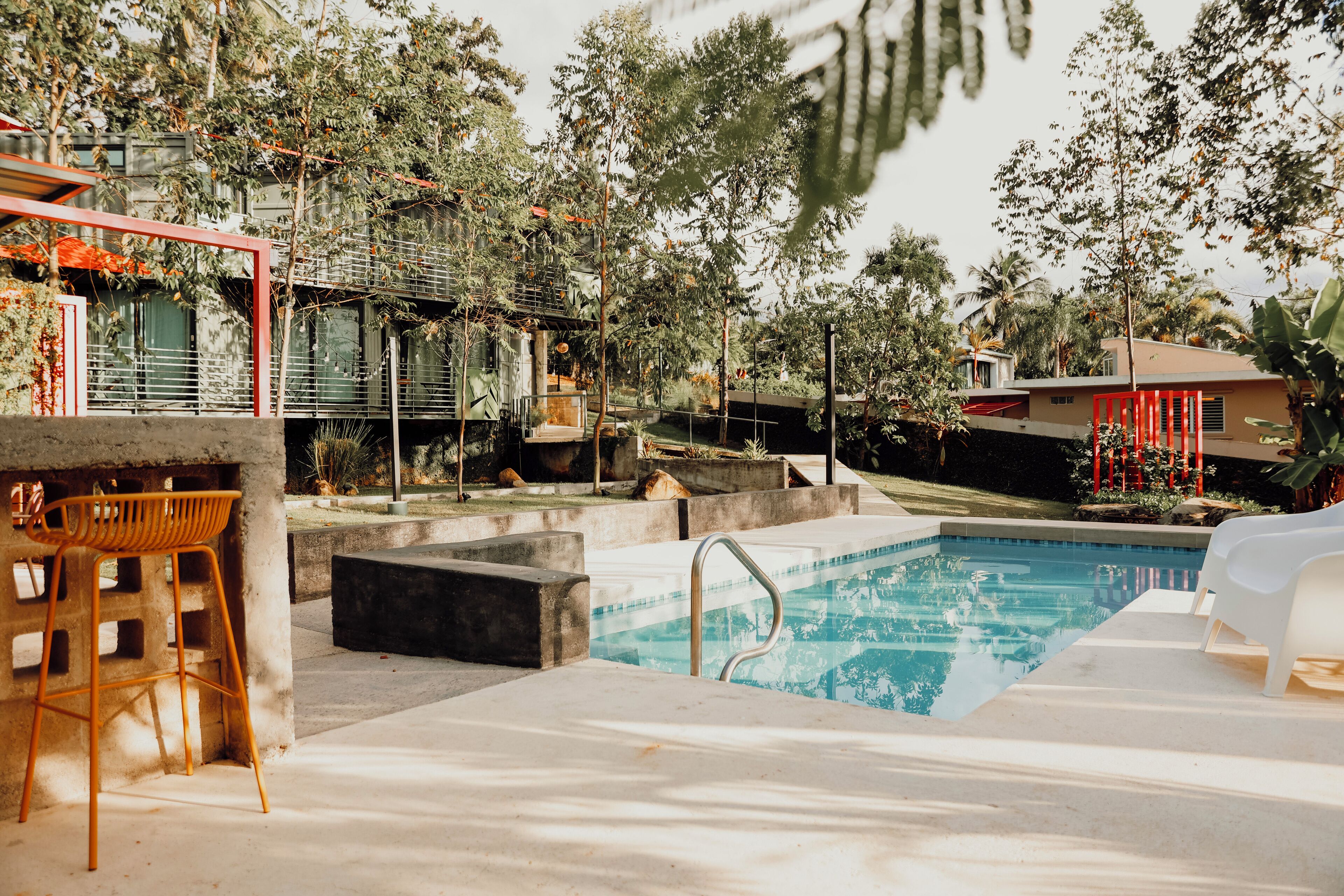 Une piscine dotée d’un bord en béton, entourée de verdure et bordée d’une structure rouge.