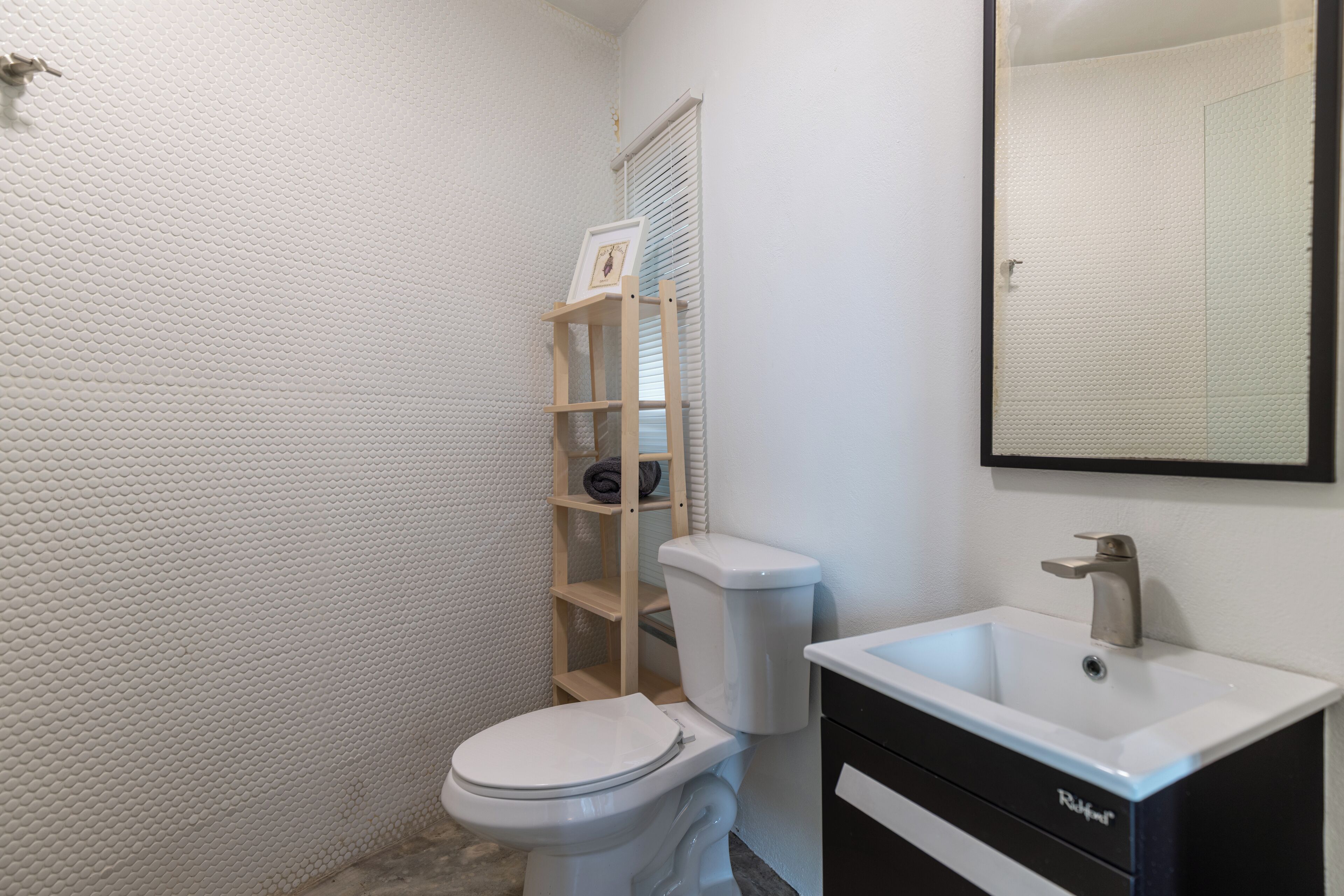 Un baño con pared blanca texturizada, un inodoro blanco, un estante de madera con toallas, un lavabo blanco con un mueble negro y un espejo sobre el lavabo.