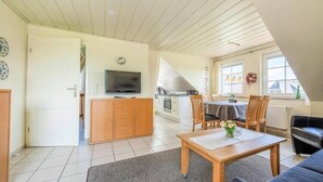 A spacious living area with a wooden table, a sofa, and a TV in the background. - Ferienwohnung im OG in Friedrichkoog-Spitze (Friedrichskoog-Spitze)
